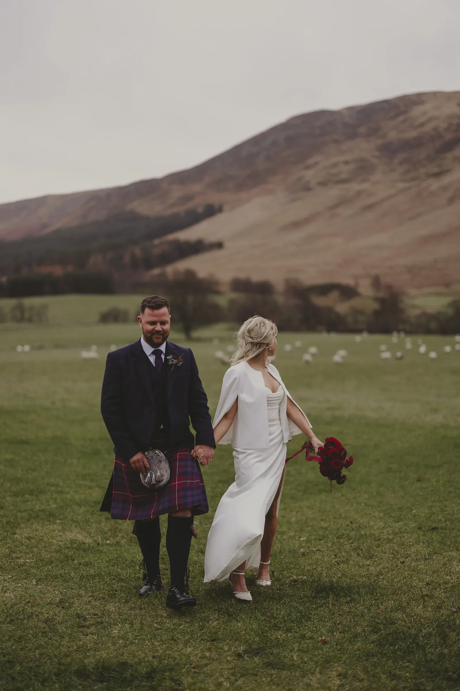Katie and Kevin walk hand-in-hand through the glen with the beautiful Scottish scenery and sheep visible in the background.