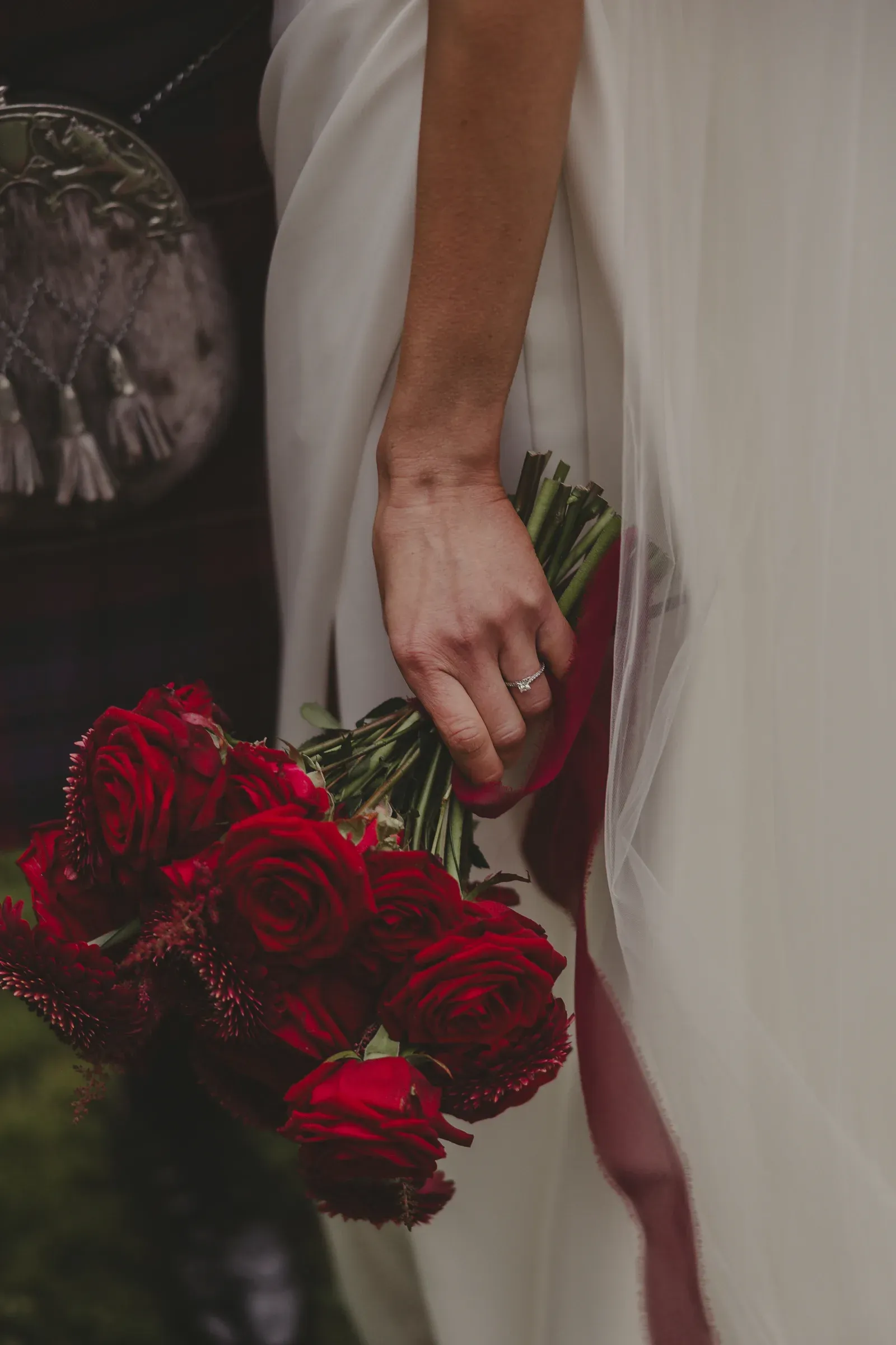 A close-up detail shot featuring a bride in a white dress holding a bouquet of red roses alongside a groom in a sporran.