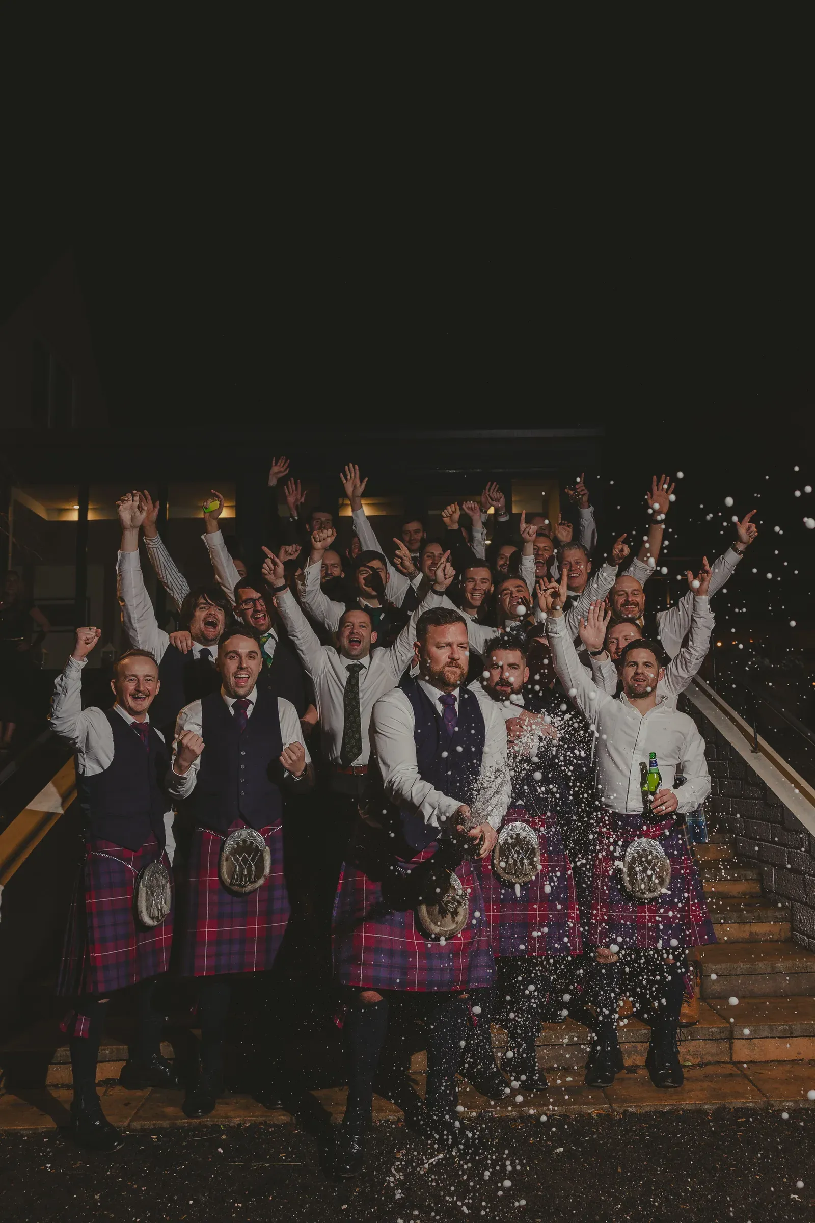 A large group of men in kilts celebrate outside the wedding venue by popping champagne and cheering.
