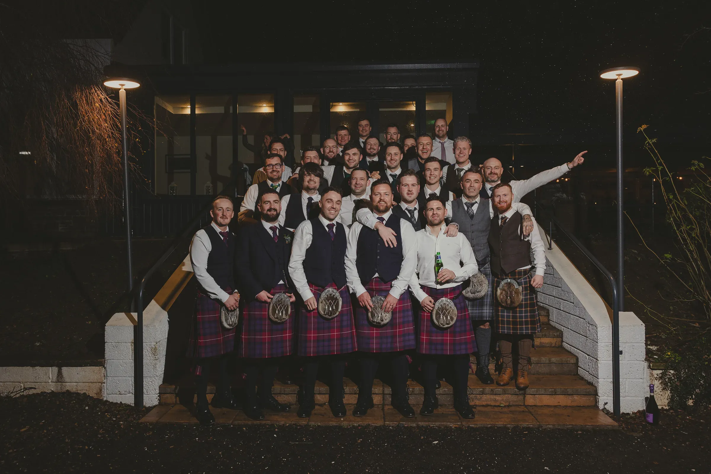 A large group of men from the wedding party, many wearing traditional kilts, pose together outside the front door of the venue during the evening reception.