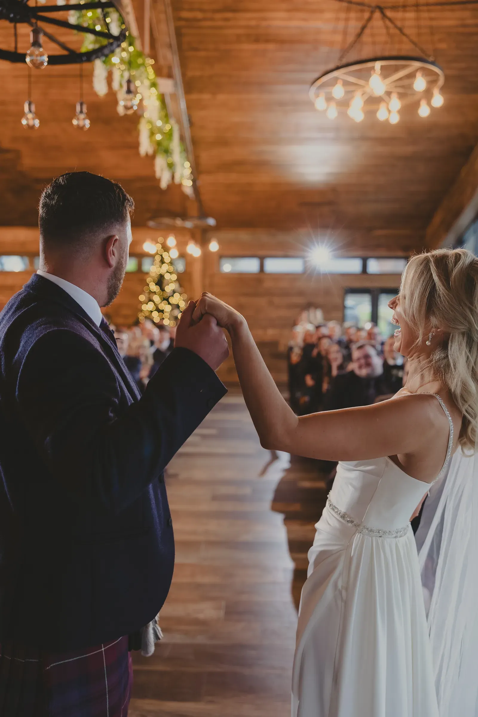 Bride and groom Kevin and Katie share a joyful moment holding hands inside the barn with their wedding party visible in the background.