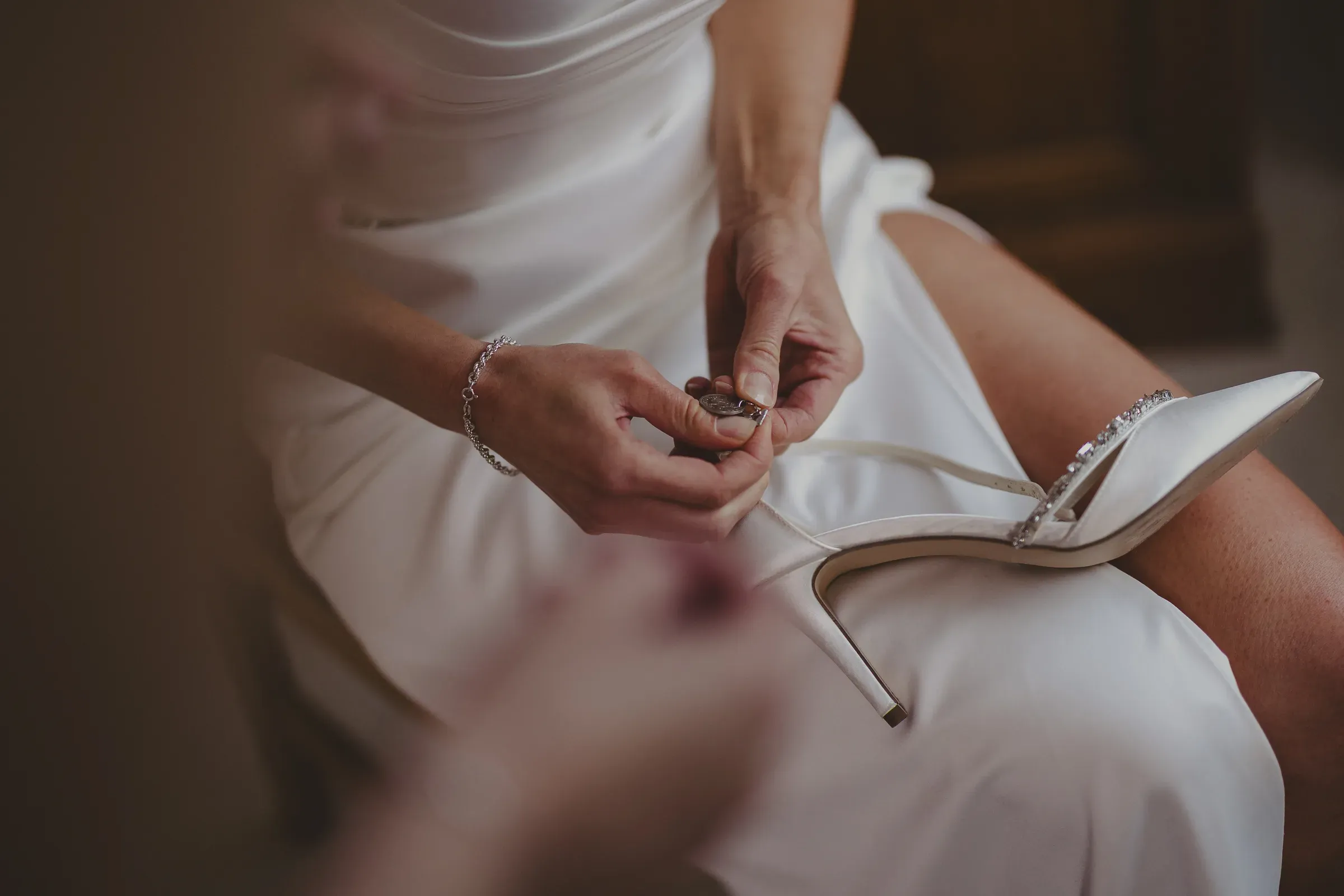 A close-up staging shot of a bride holding her wedding shoe, highlighting her bracelet and the aesthetic details of her bridal attire.