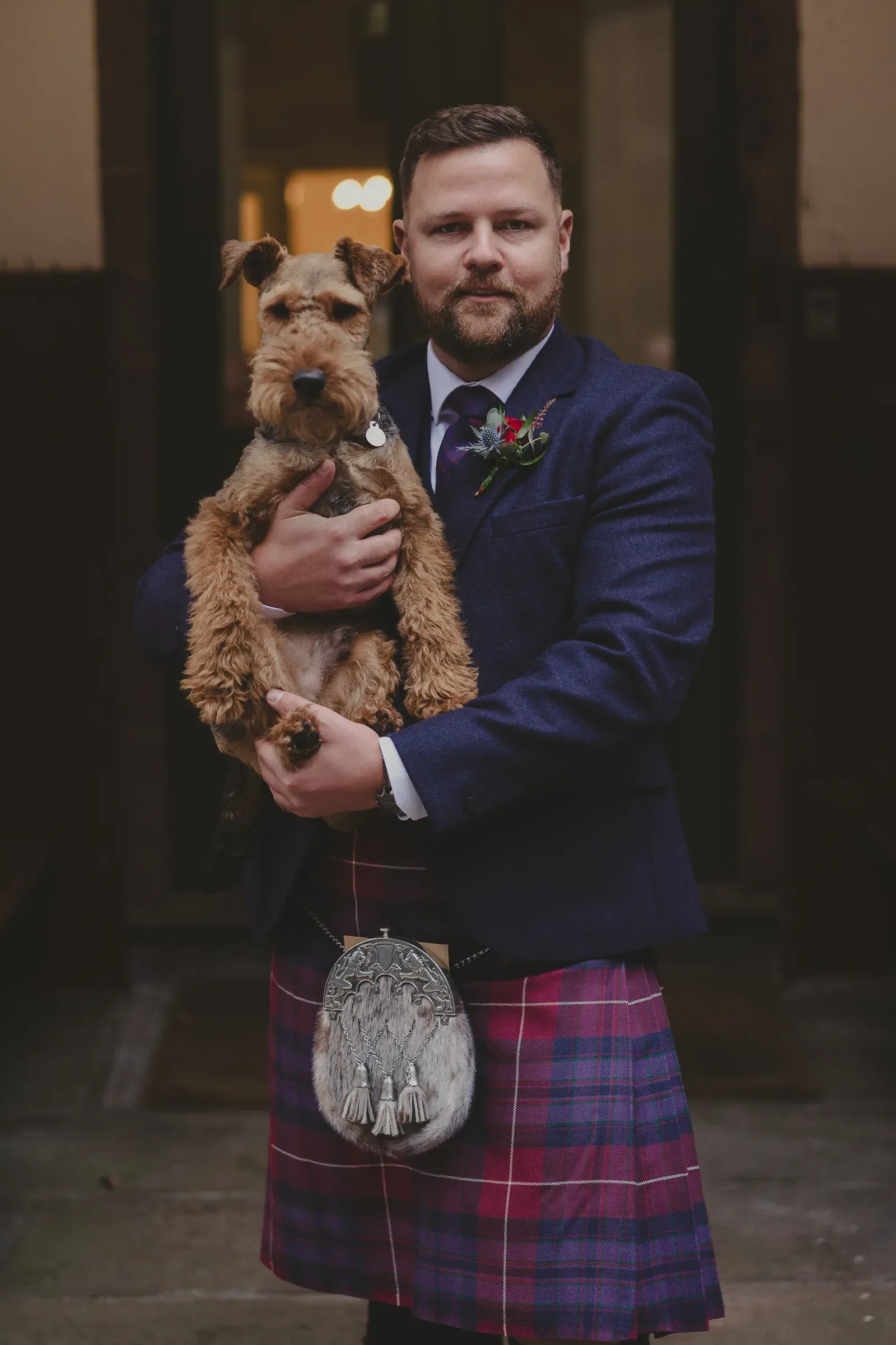 Kevin holds his dog, who is wearing a matching tartan accessory, during his wedding day.