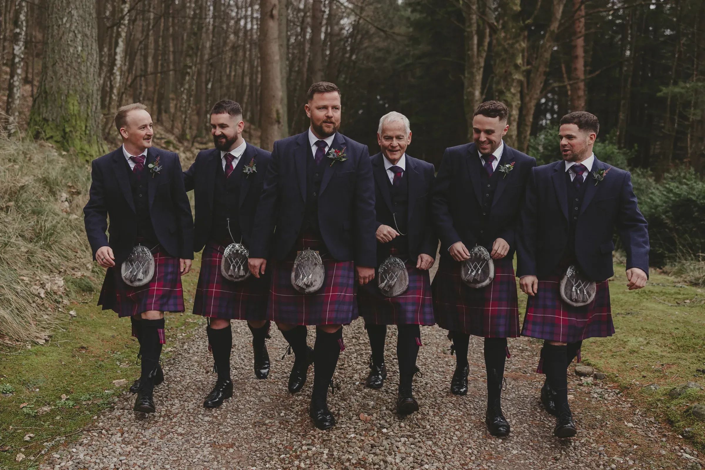 The groom party for Katie and Kevin's wedding walks together along a wooded path near the hotel, all wearing matching kilts.