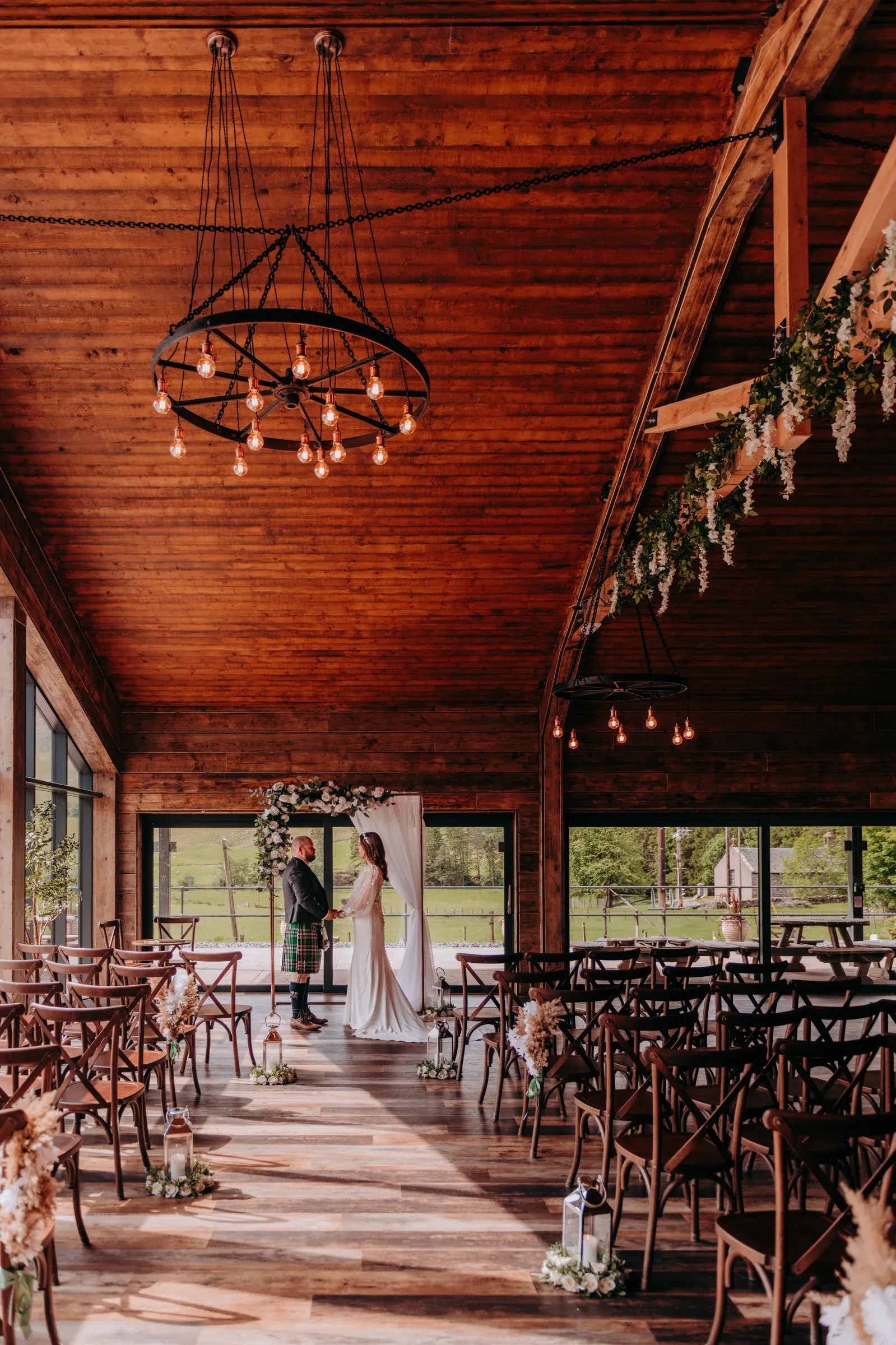 Sam and Cam stand at the altar inside the barn, captured from a distance to showcase the venue's wooden architecture and overhead lighting.