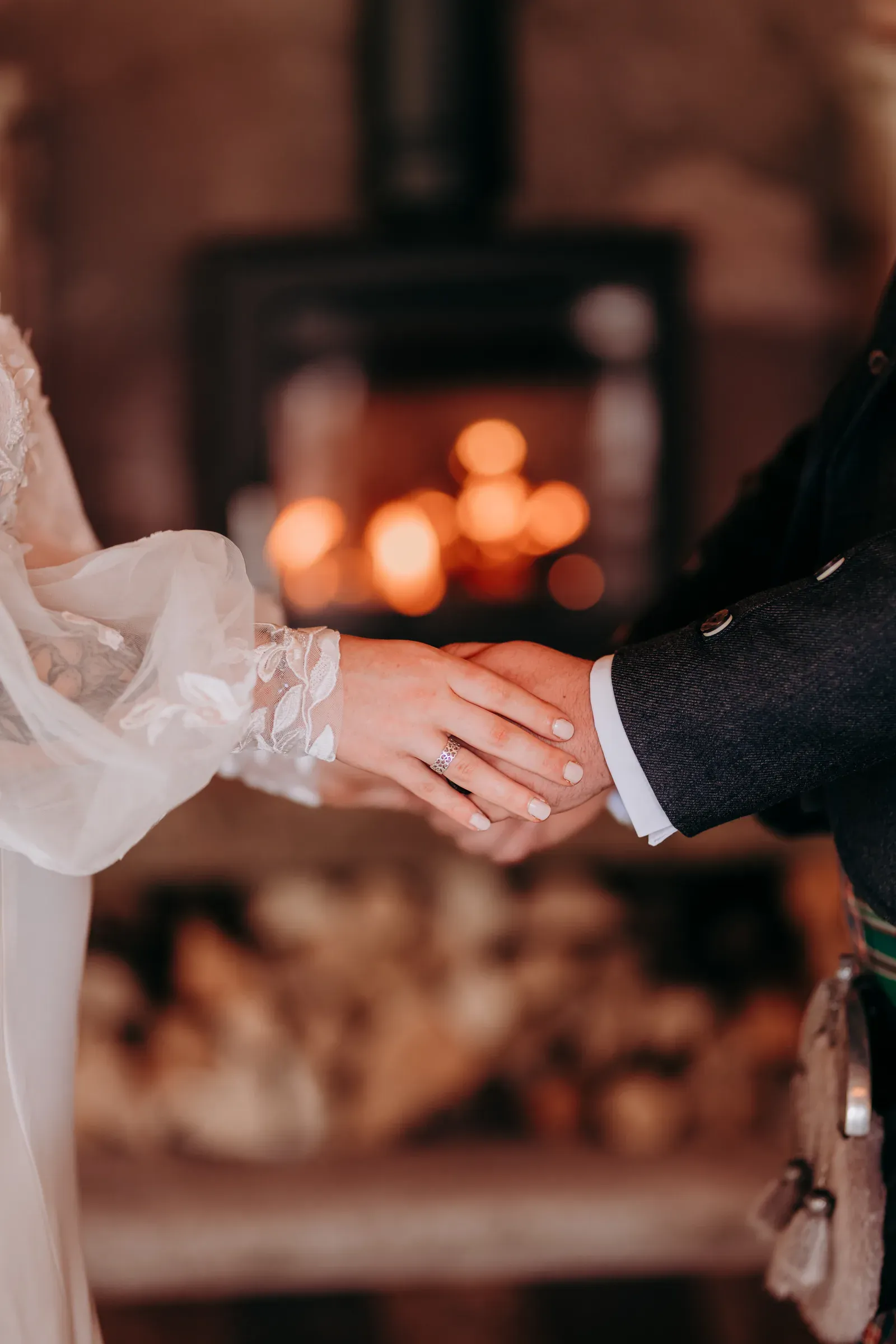 Sam and Cam hold hands in front of a fireplace, highlighting the wedding ring against the natural fire glow.