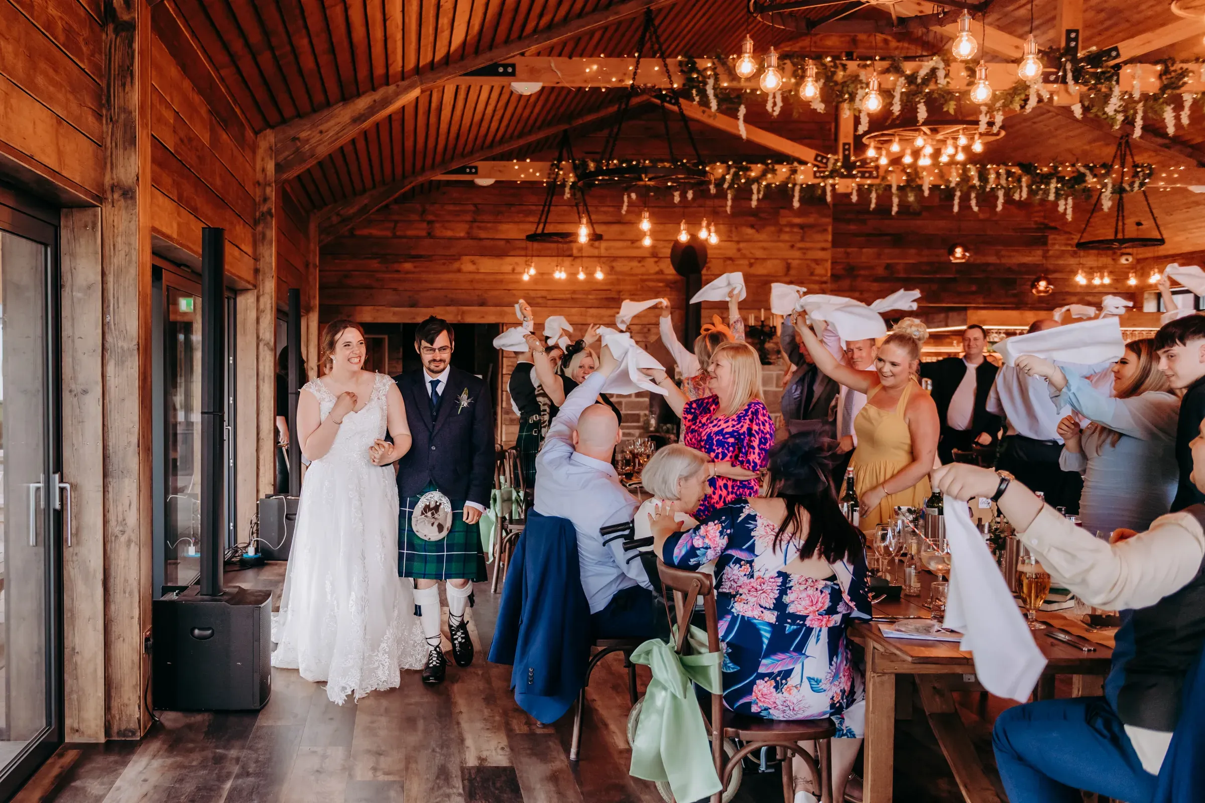 Heather and Louis walk through the barn at Glenclova as their wedding party celebrates by waving napkins during dinner.