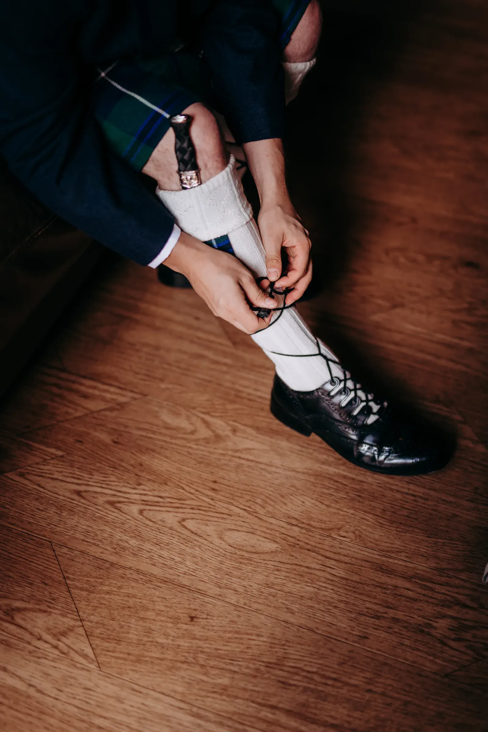 A groomsman in traditional Scottish attire ties his shoelaces in a staged preparation shot.