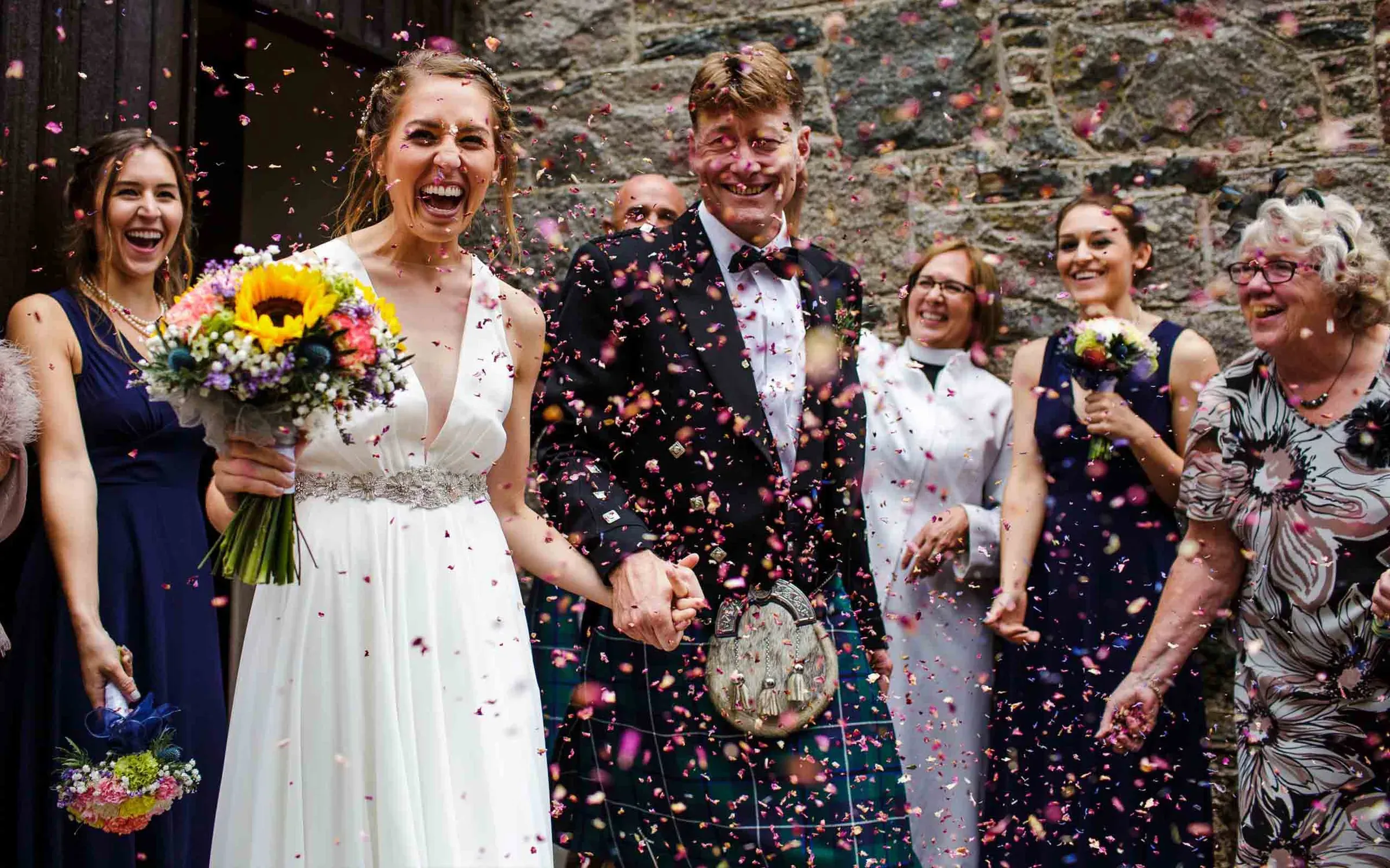A happy bride and groom walk through a shower of confetti surrounded by smiling wedding guests.
