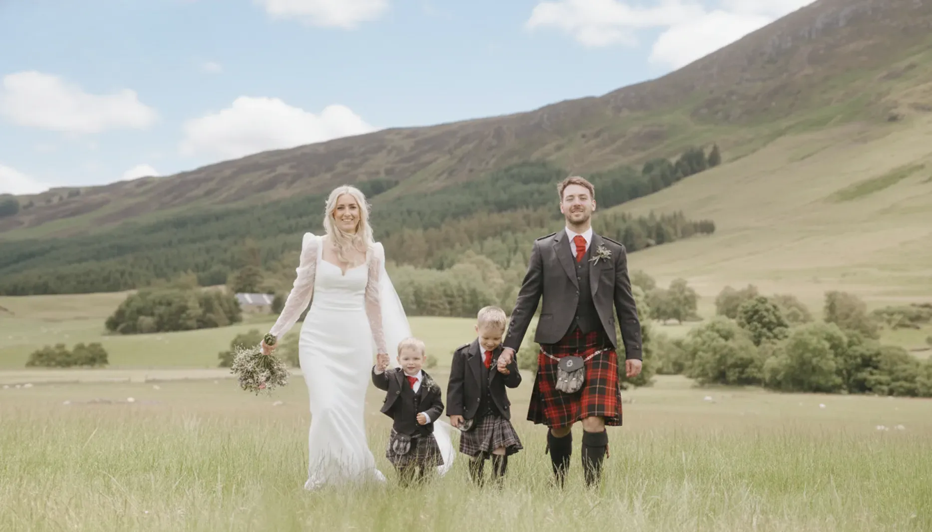 A bride and groom walk through the glen holding hands with two young children in a family-focused wedding portrait.