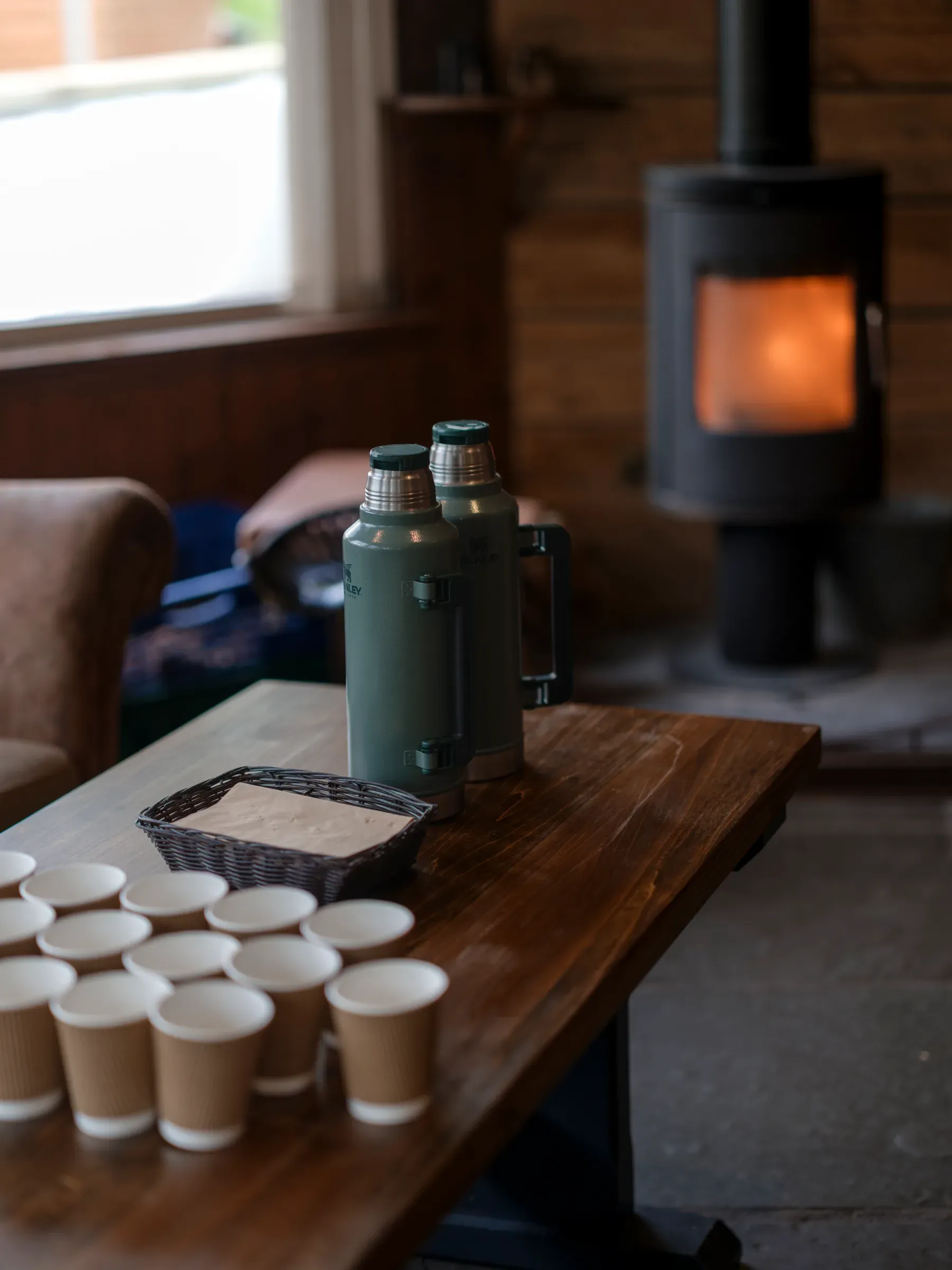 A warm and cozy hot chocolate and coffee station set up on a wooden table with a glowing fireplace in the background.