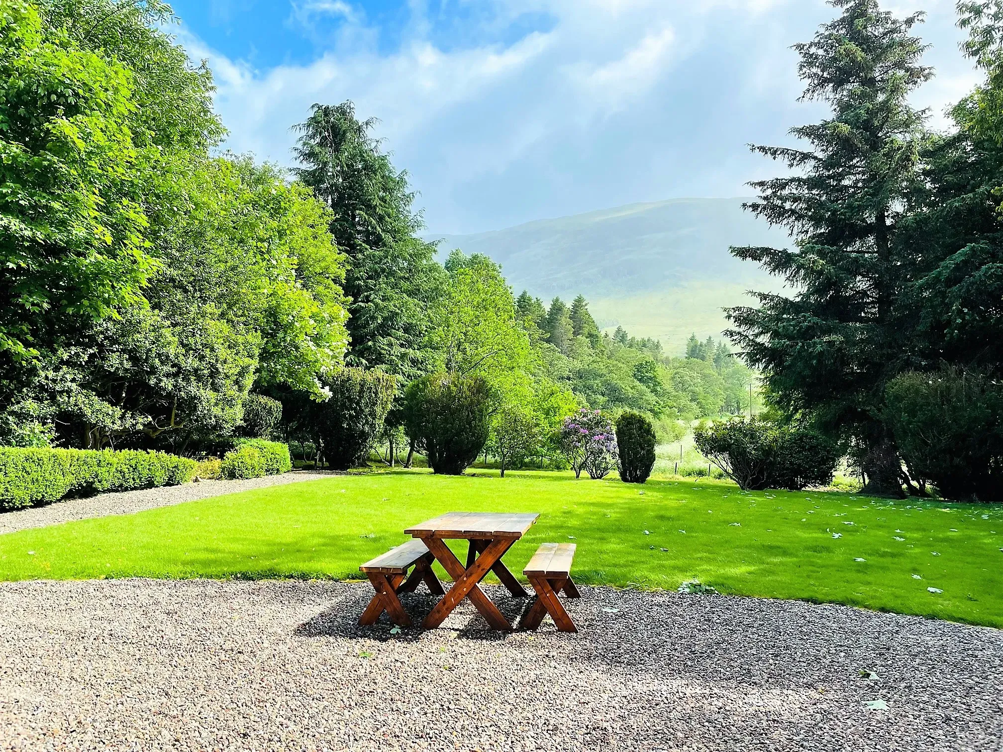 A wooden picnic bench sits on a gravel area in front of a lush green lawn with a cottage and scenic glen landscape in the background.