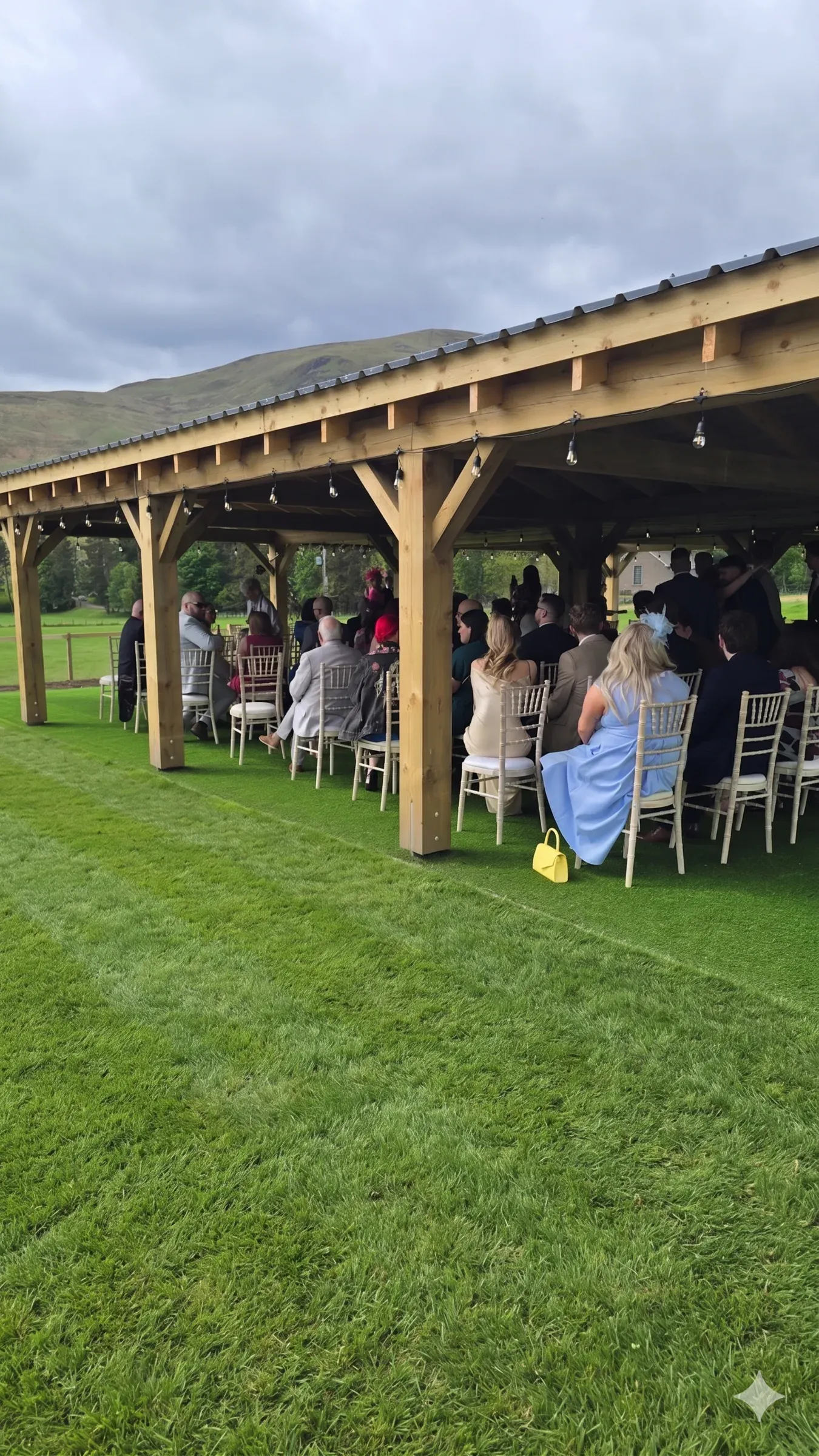 Wedding guests seated in the meadow under the outdoor structure during a ceremony.