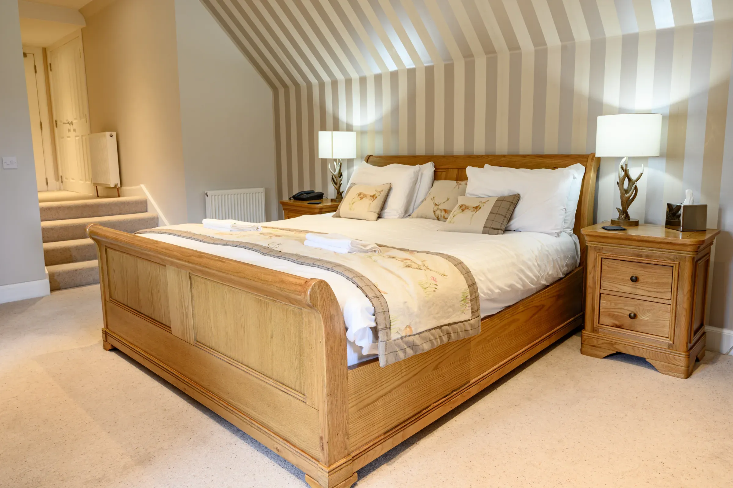 A view of a neatly made hotel room bed featuring wooden furniture and striped wallpaper.
