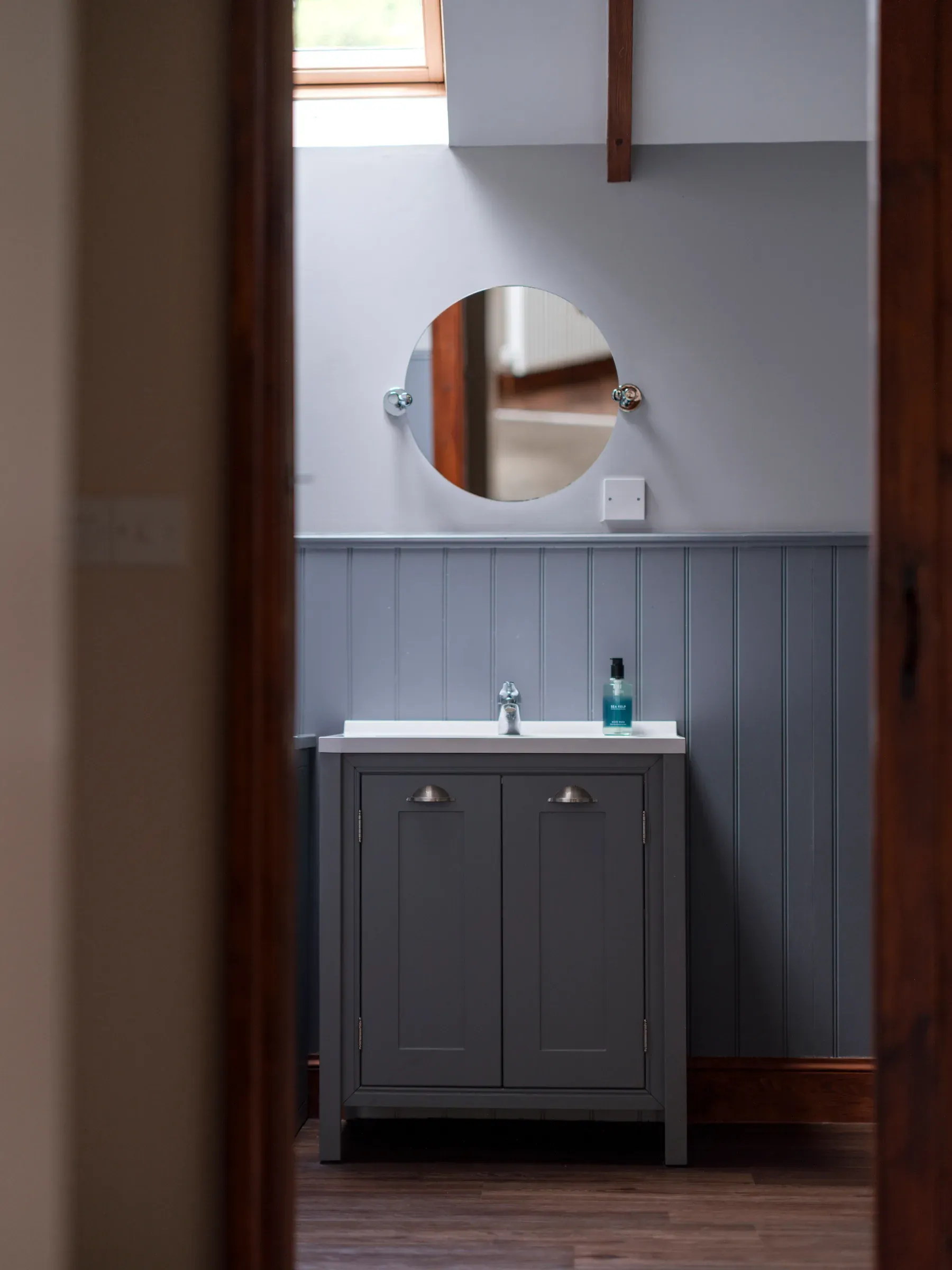 A view into a modern bathroom featuring a grey vanity unit, circular mirror, and wood-panelled walls.