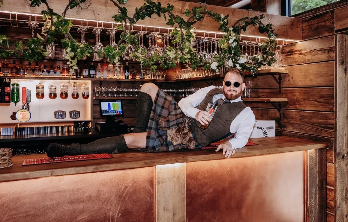 Callum lying along the barn bar in his kilt with a pint, after the "Where the hell is my husband?" call-in